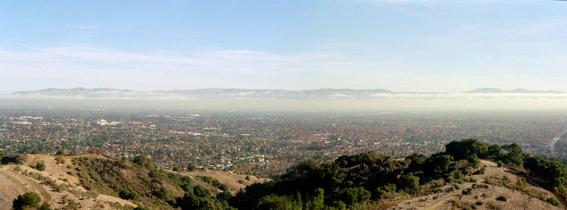 [Two images stitched together showing even more of the hilltops in the foreground and more of the valley below and the mountains in the distance. While there is a haze of white over the valley, the air is not as thick and thus more detail is visible than of the other image with the haze. There are strings of white clouds just below the tops of the mountains as well as wispy ones higher in the sky. There are dots of red and green in the valley as some trees have changed color while others have not.]